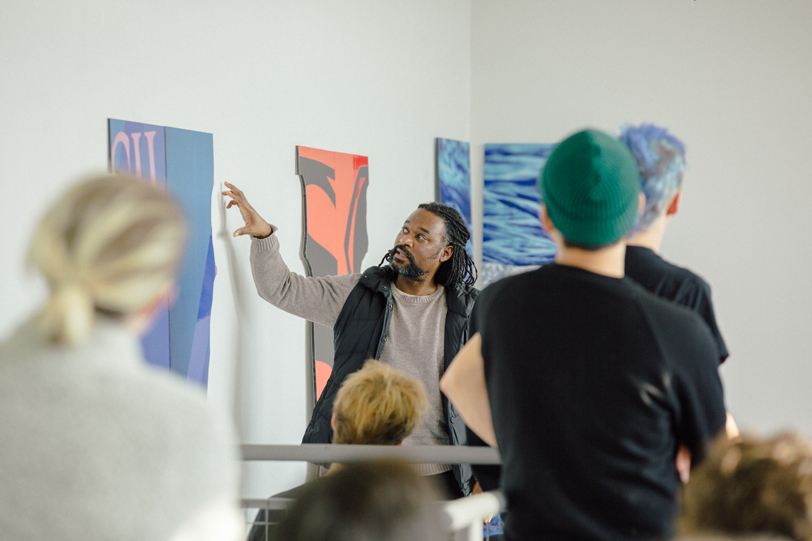 A teacher points describes a painting on a wall to a group of students