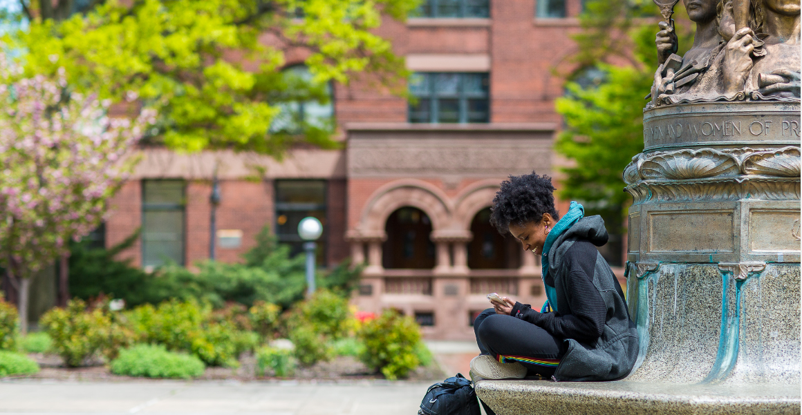 A student looks at her phone while sitting at the steps of a statue.