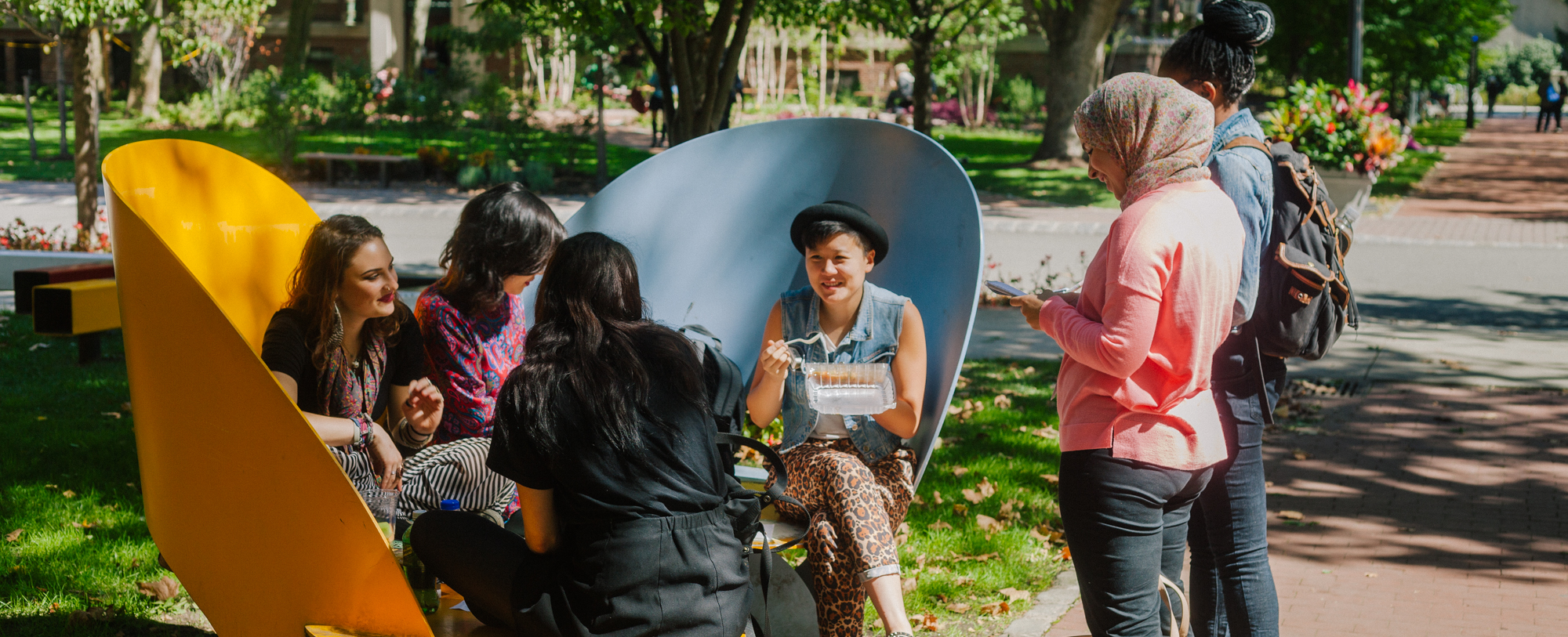 Six students sit and stand in a circle as they eat lunch in a park.