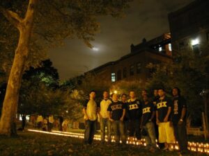 A group of Fraternity members stand in a park at night with a trail lit by candles on the ground in the foreground.