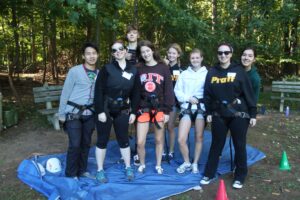 A group of students look at the camera while wearing zip-lining gear and standing on a blue tarp. There is a wooded are in the background.