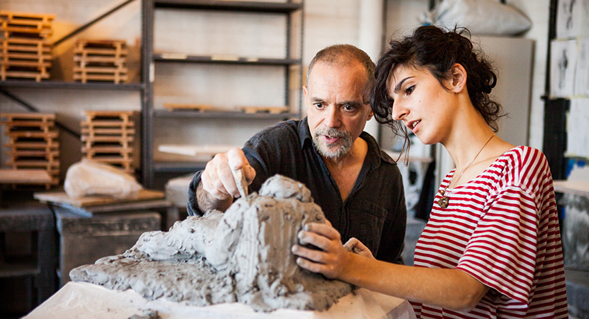 A student observes a professor displaying sculpture techniques by pointing at a pile of clay.