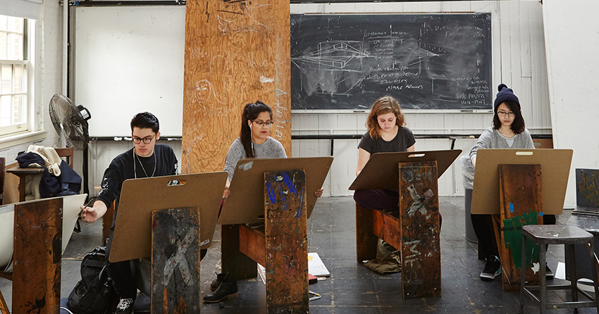 Four students sit in front of stencils in an art studio as they work.