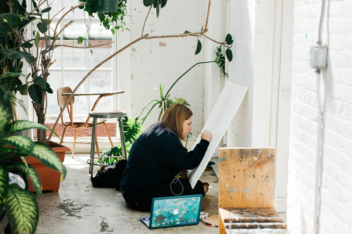 A student draws on a stencil while sitting on the ground in a well lit rooms with plants and art supplies.