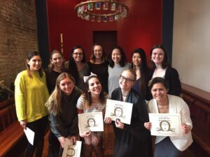 Soririty members hold diplomas while taking a group photo inside a red room with an intricate chandelier hovering overhead.