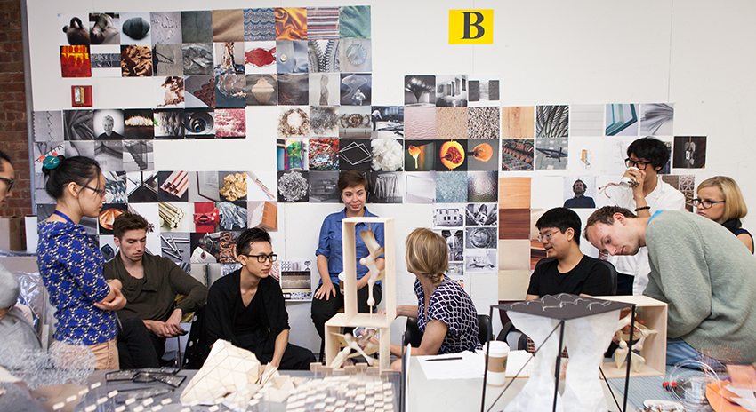 A group of students sit around their professor in front of a wall covered with photos and graphic design images.