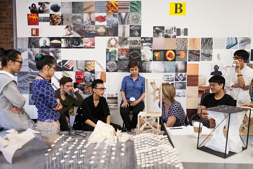 A group of students sit in a circle discussing with their professor with photographs in the background.