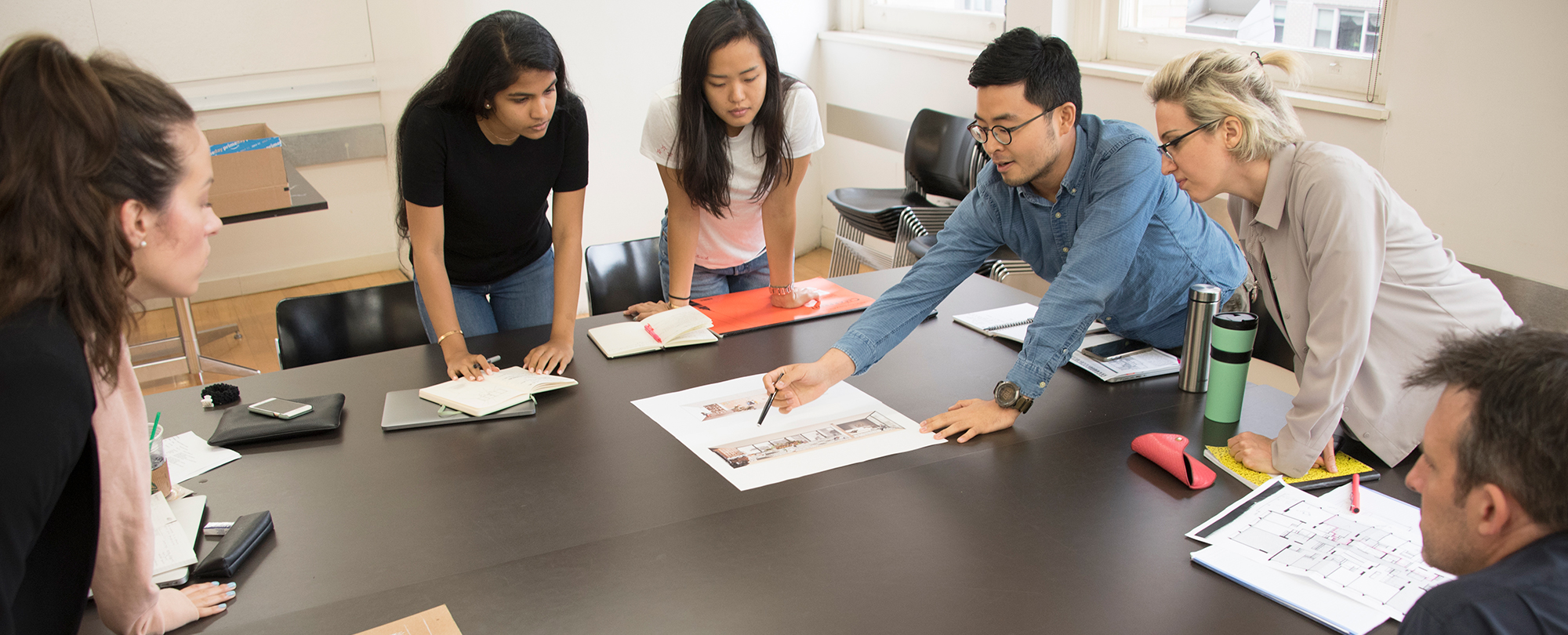 A group of people stand around a table with schematics as they discuss them.