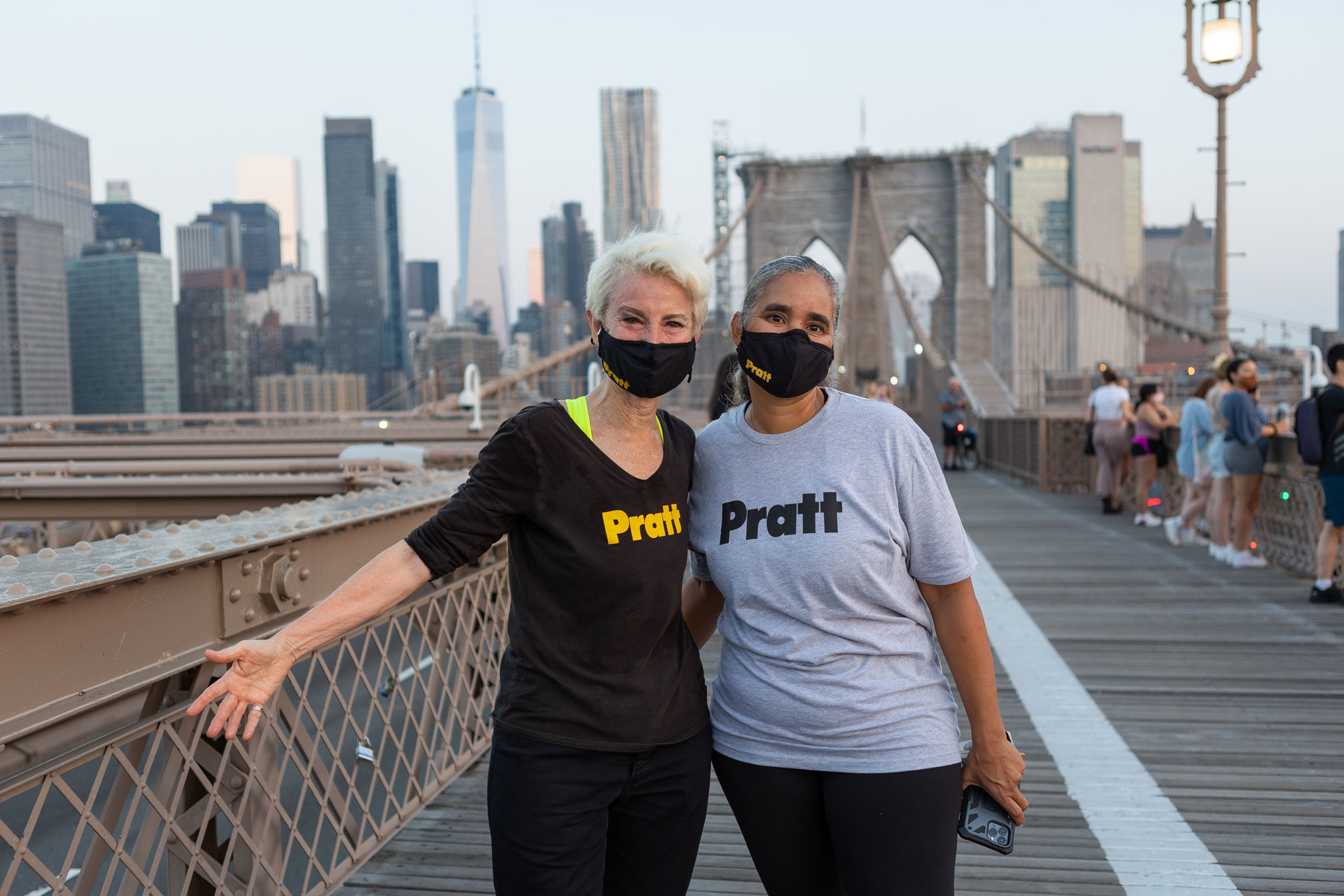 President Bronet and another staff member are on the Brooklyn Bridge looking at the camera. They are wearing masks and t-shirts with the Pratt logo. The New York City skyline is behind them and students are looking out over the side of the bridge.