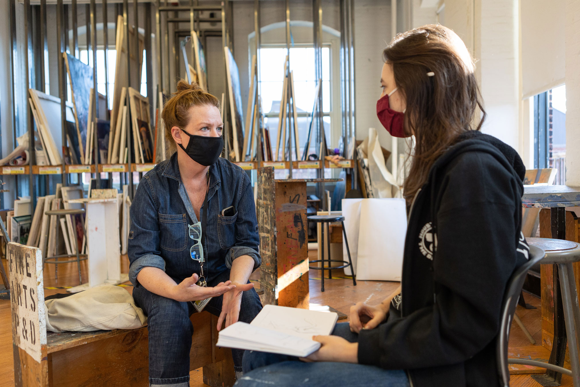 A faculty member and student are sitting in a studio space conversing. There are canvases behind them on stored on racks.