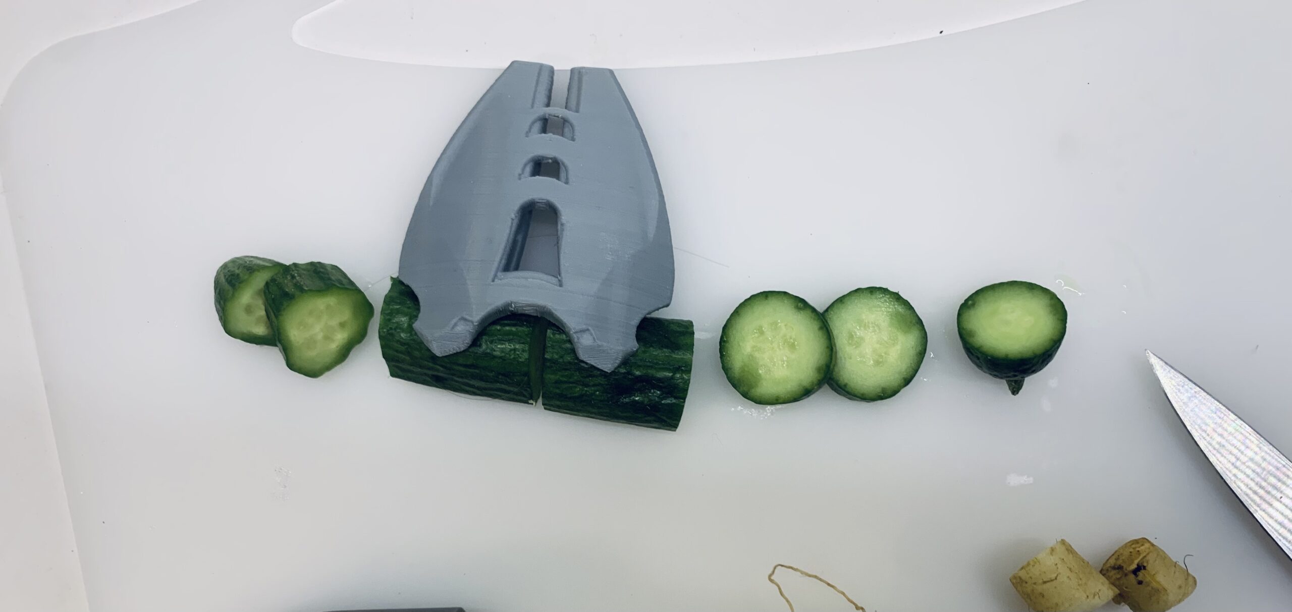 Blue attachment grips a cucumber, slices of the cucumber are on either side of the attachment, as well as a knife resting on the cutting board.