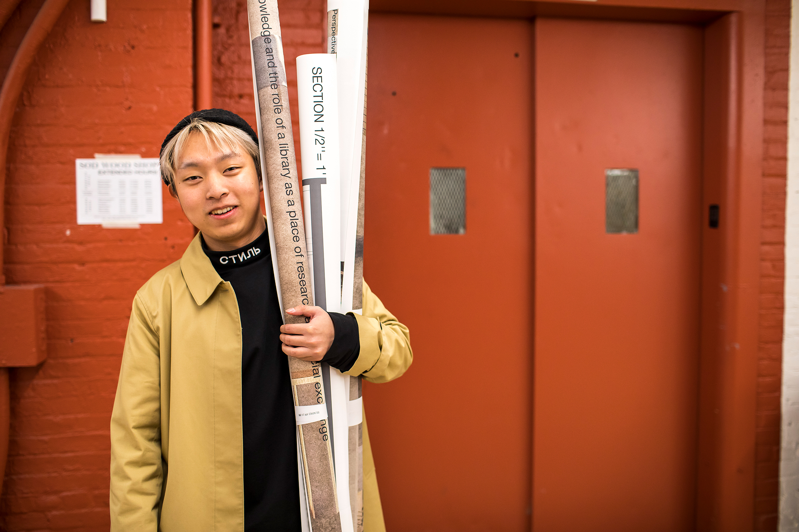 A student is smiling standing in front of a red wall and elevator. They are holding large rolls of printouts.