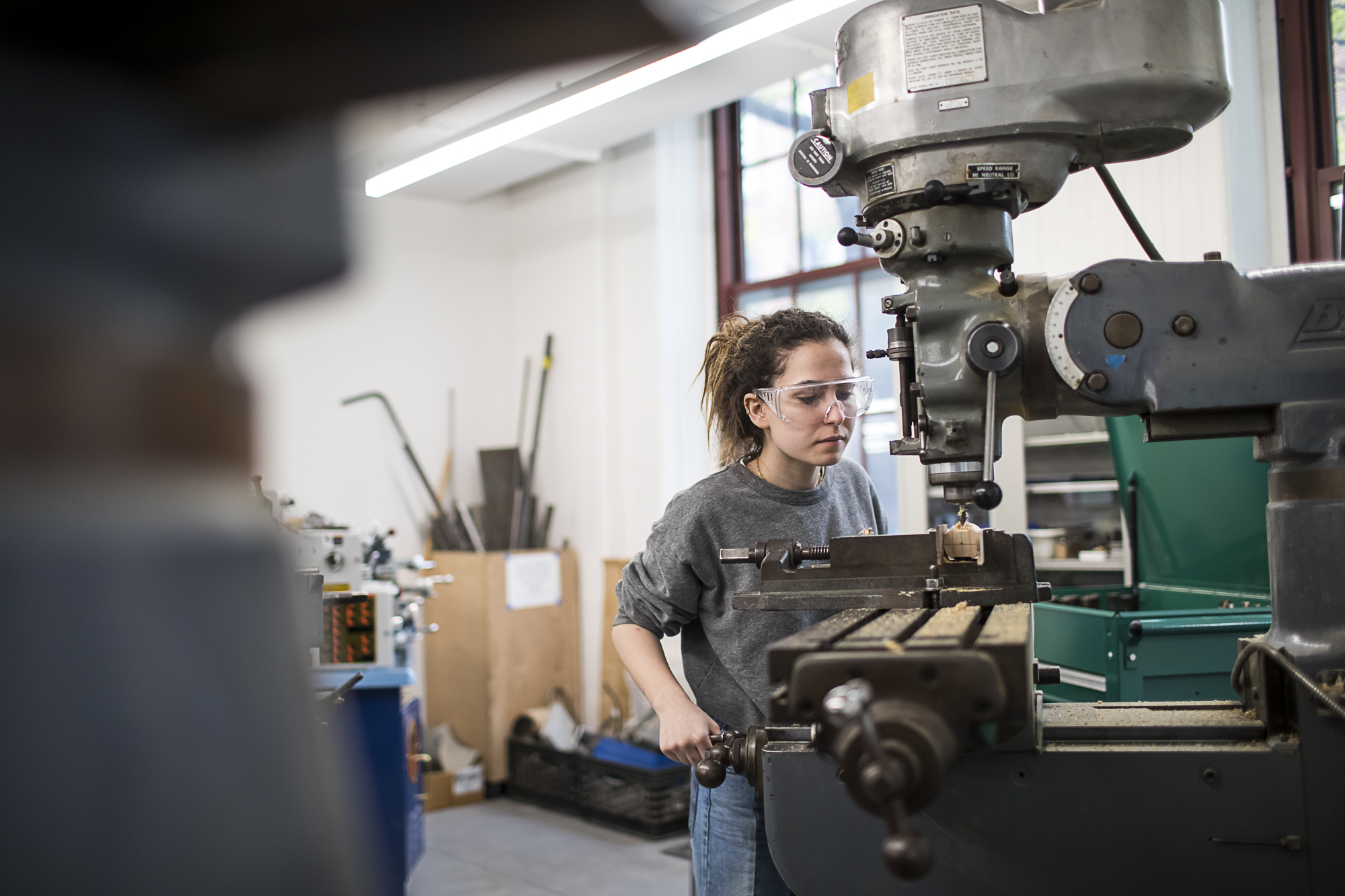 A student in a machine shop is working with a large drill. They are wearing safety goggles.