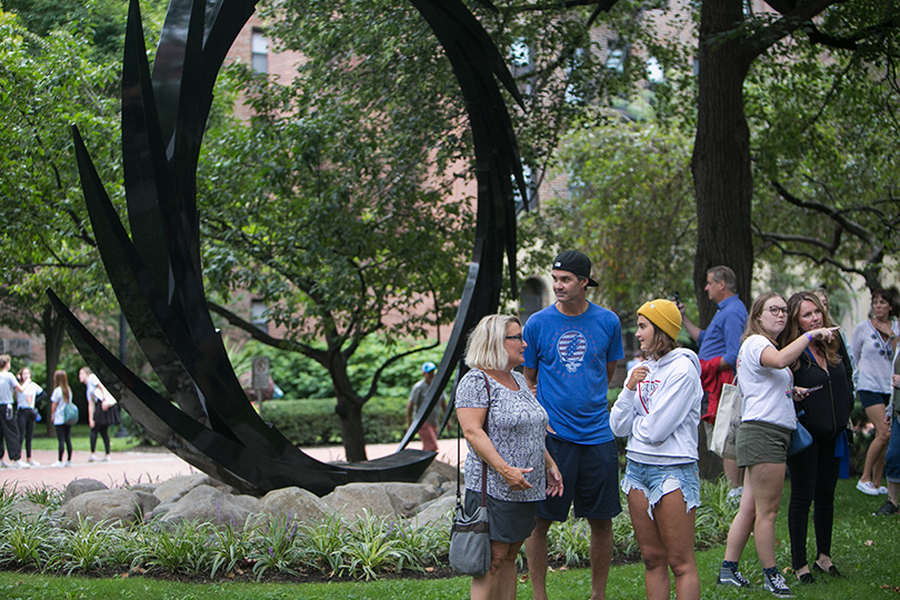 Students and parents stand in groups on campus with a large sculpture behind them.