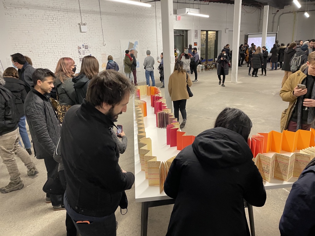 people crowd around a l-shaped table topped with accordioned papers that have drawings on them