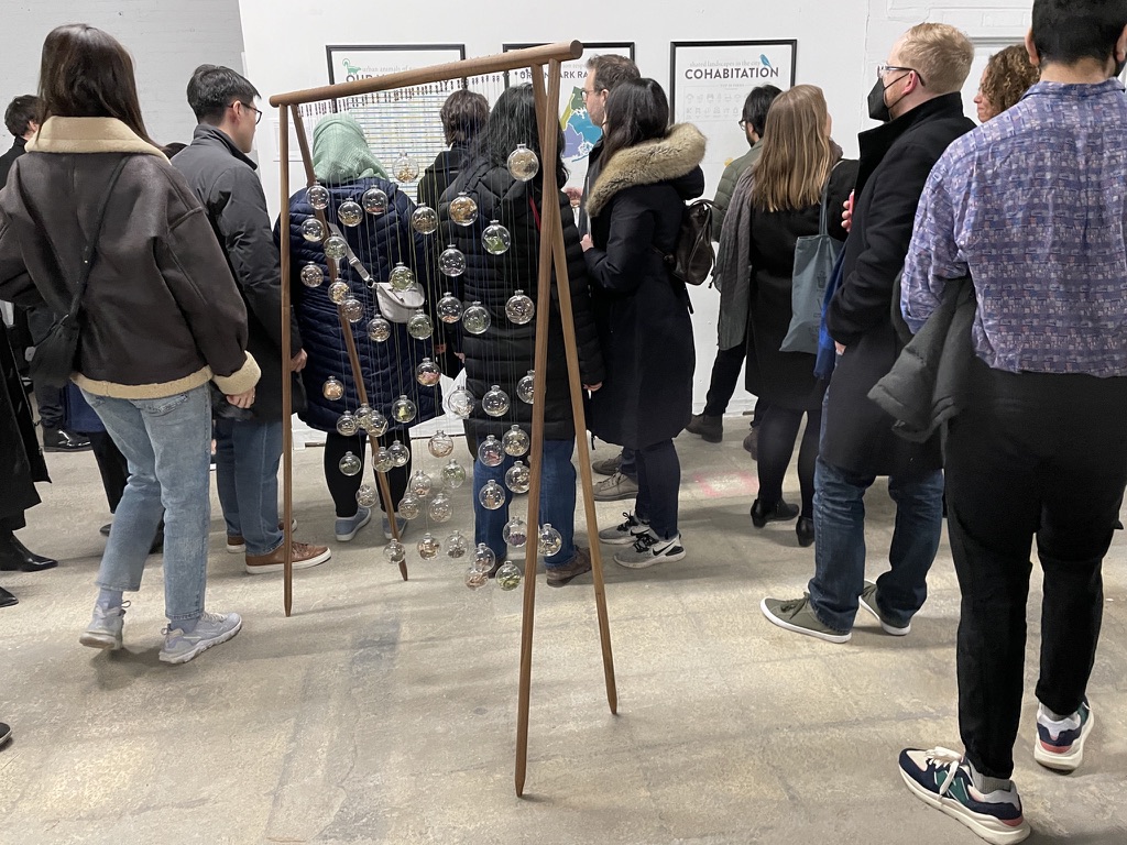 people surround a sculpture that resembles the support of a swing with glass orbs hanging from the bar.