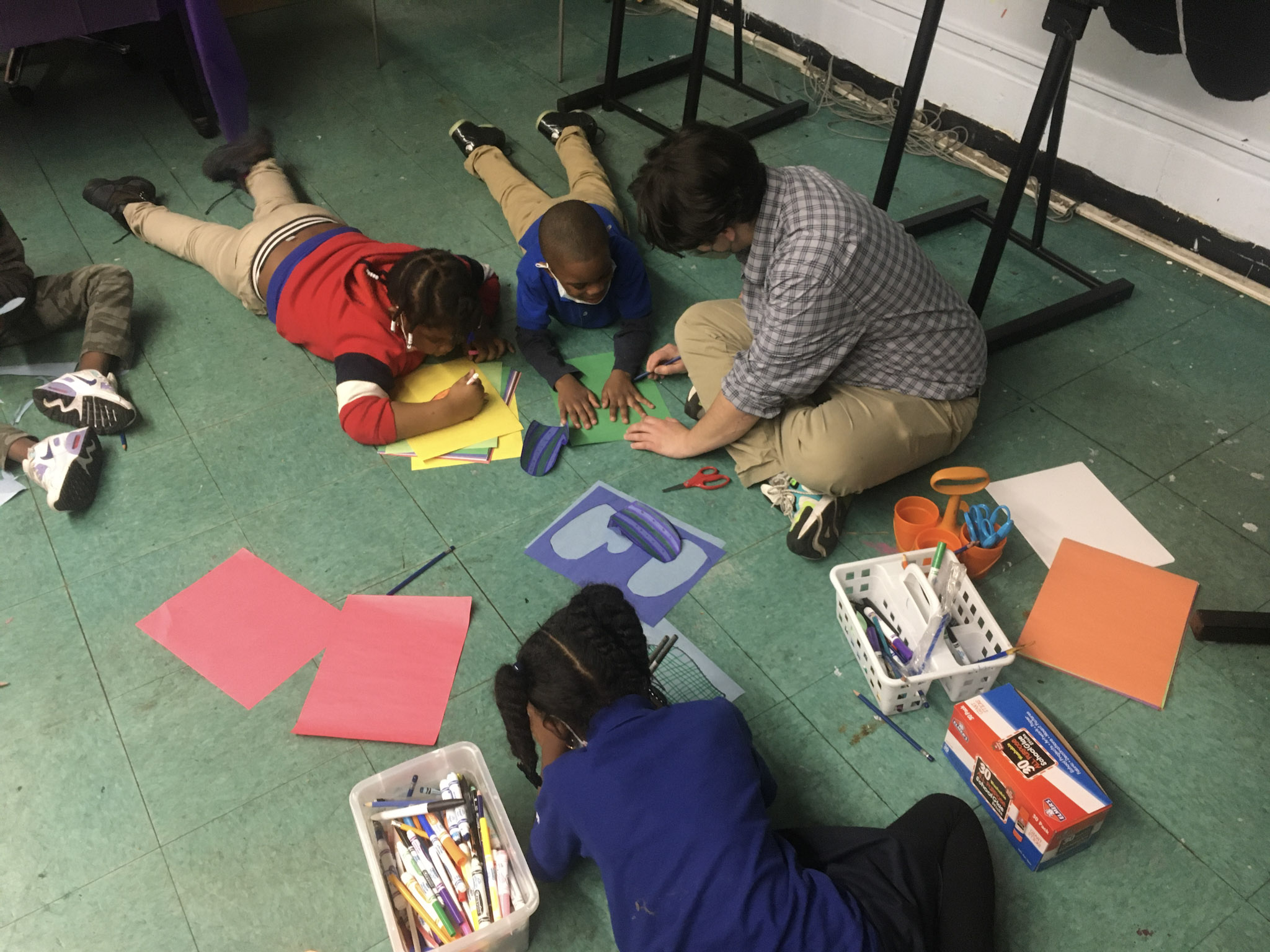 teacher instructs students as they draw and cut out paper on the floor
