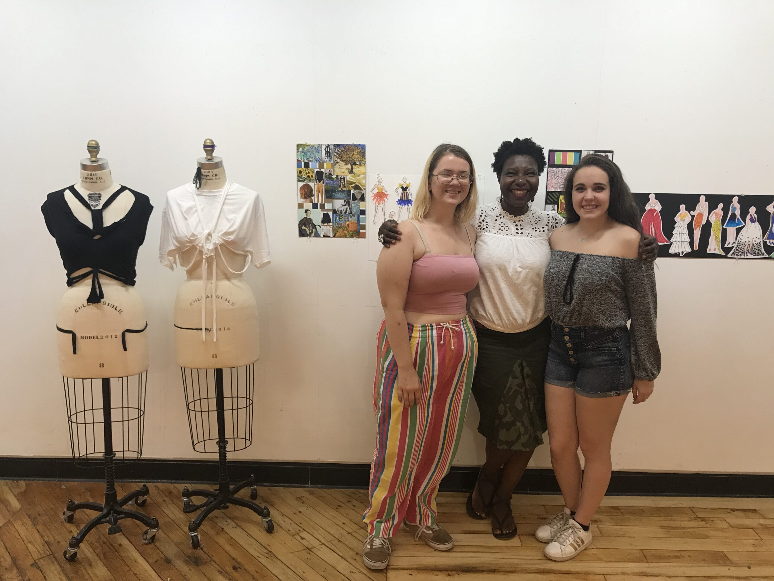Three students stands in front of their fashion design projects, with two mannequins on their right and a set of fashion sketches behind them on the wall.
