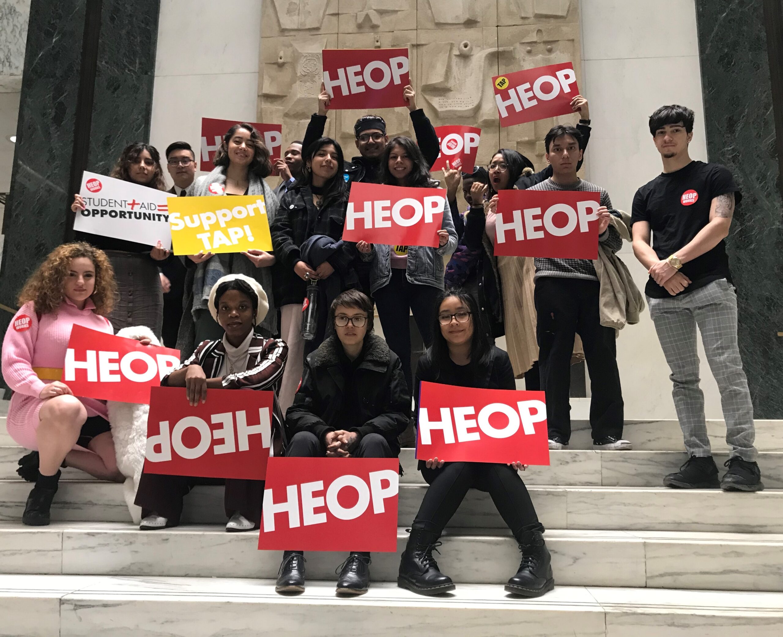 A group of students pose for a group picture atop a set of marble stairs. They are carrying red banners with the word 