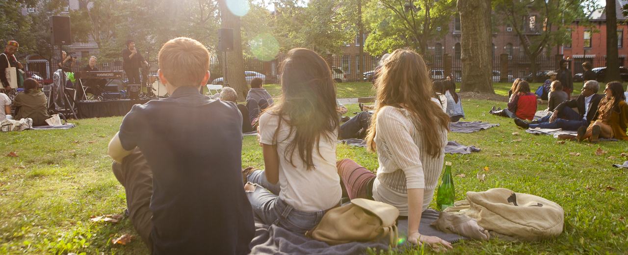 Three students sit in lawn facing a group of musicians performing for a crowd.