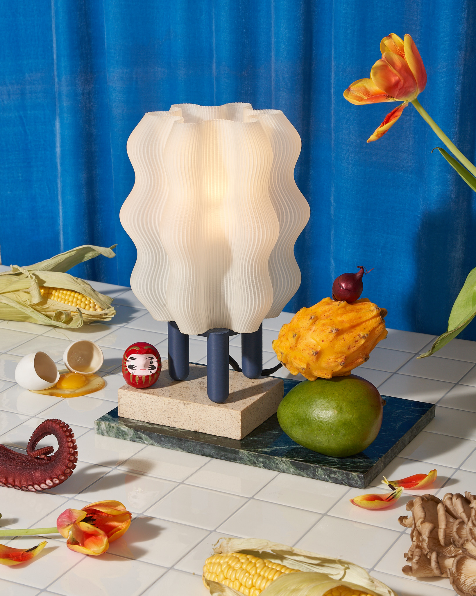 A white semi-opaque lamp with wavy, ridged sides sits on a base with three cylindrical legs. the lamp is on a piece of marble on a tiled table which also has other objects scattered around it: an ear of corn, a broken egg, fruit, the tentacle of an octopus, flowers, and mushrooms.