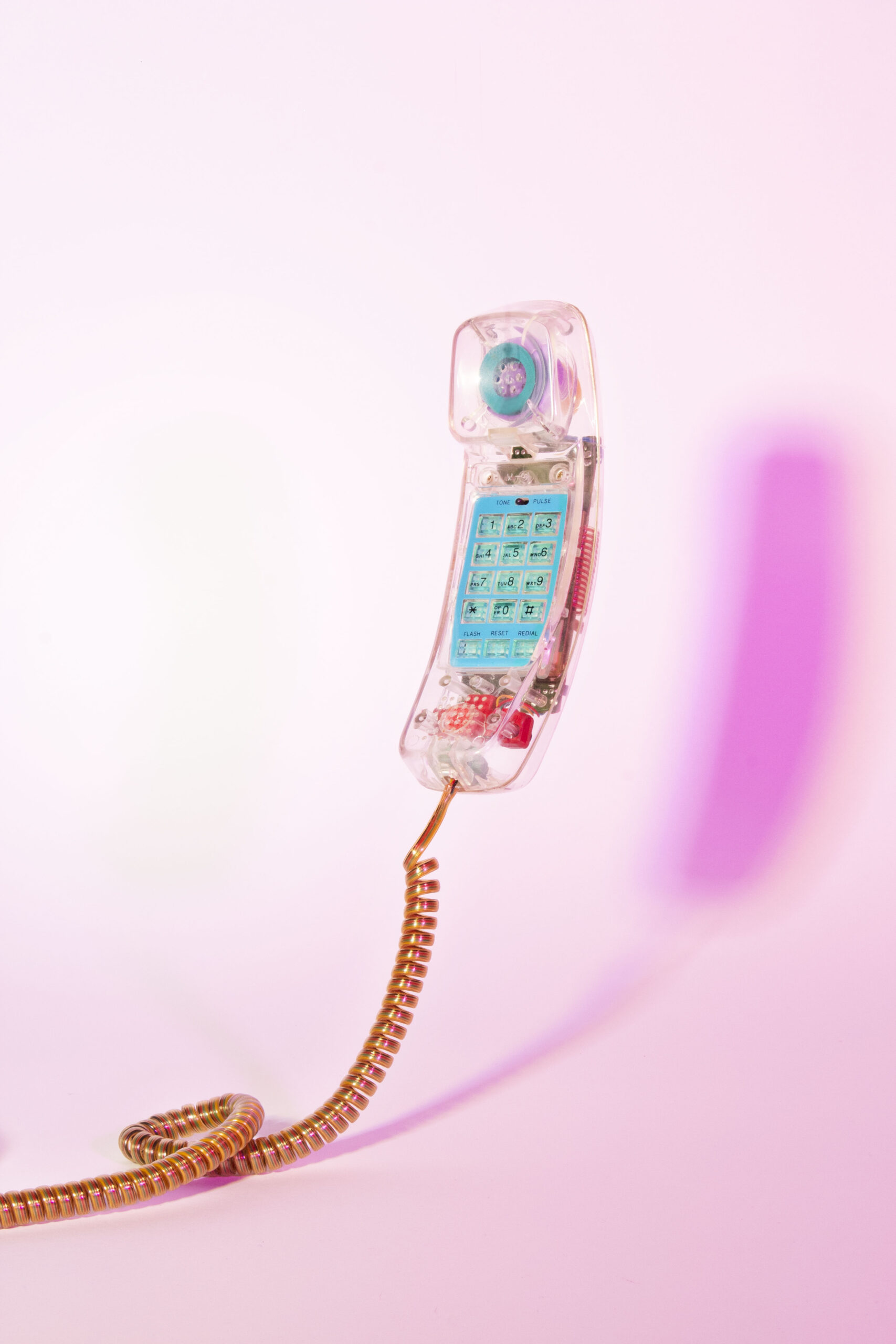 A photograph of a translucent telephone suspended in air in front of a pink to white gradient background. the shadow of the phone is on the background and a tightly coiled cable descends from the bottom of the phone and snakes to the left and out of frame.