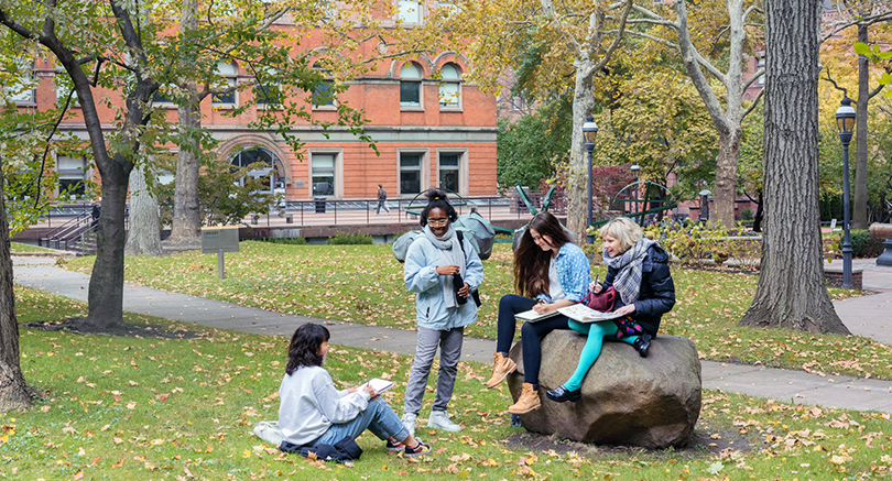 Student sit on and stand around a large boulder as they chat.