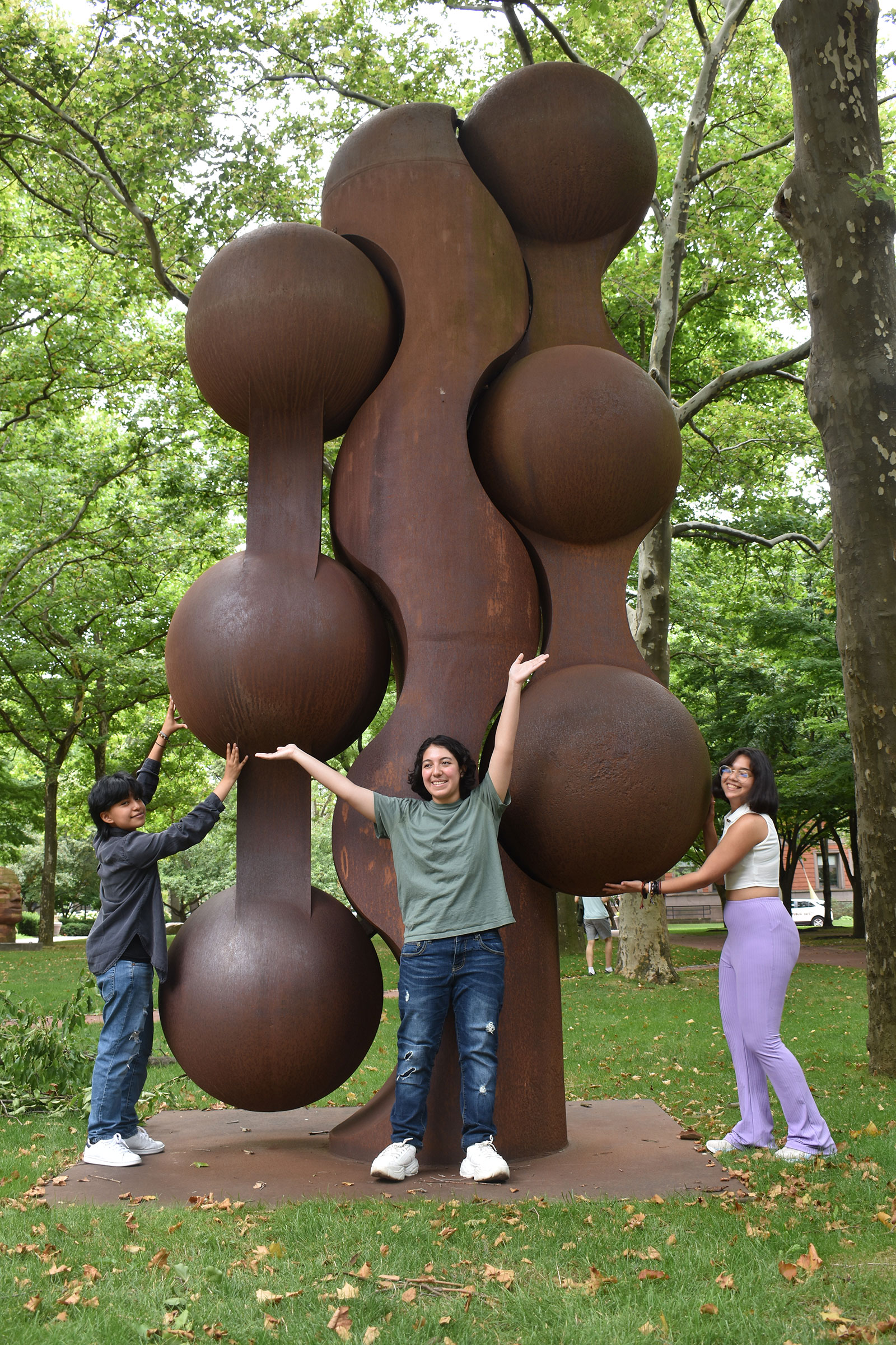 Three students pose with a large metal sculpture in a lush park.