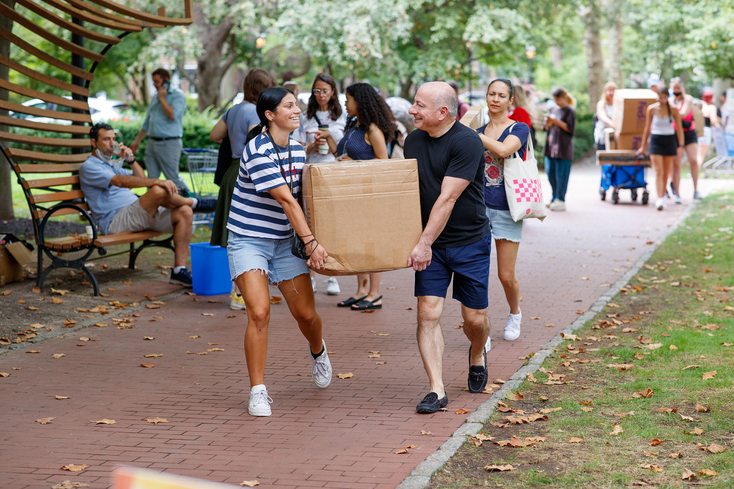 a student and their parent carries a box along a pathway through campus smiling at each other.