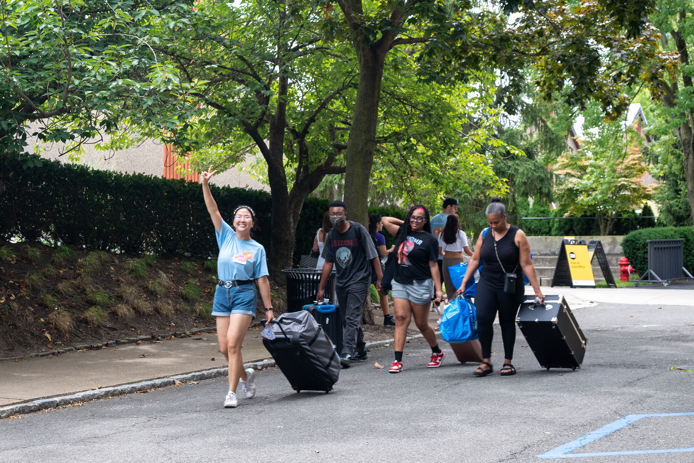 students wheeling luggage and bags through campus. One student raises her hand in the air giving the peace sign.