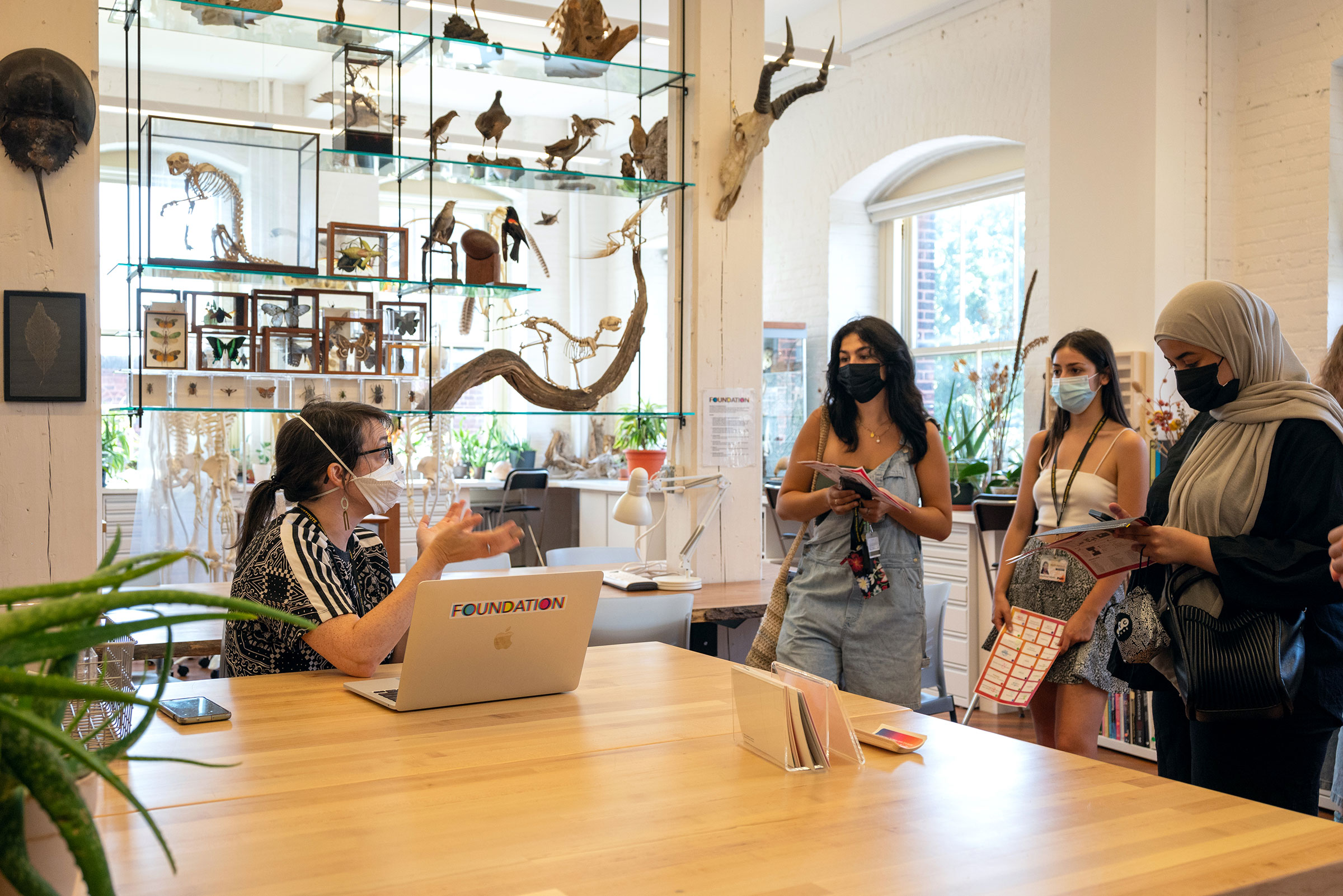 three students are listening to a fourth student as they explain about the Foundations Lab. The room is a brightly lit space with sketching and figure models arranged on glass shelves