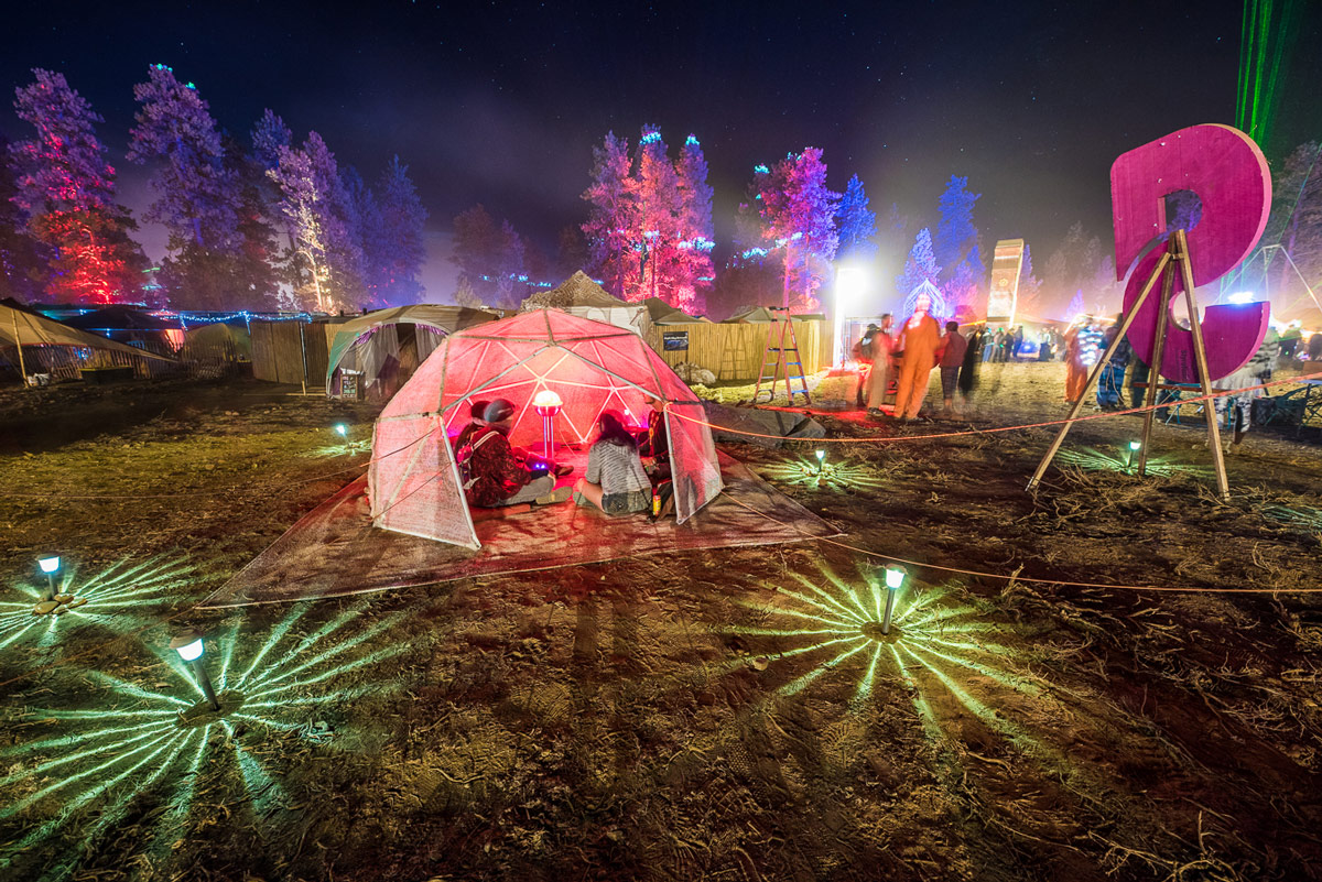 People sitting in a colorful, small dome within festival grounds that are lit up at night.