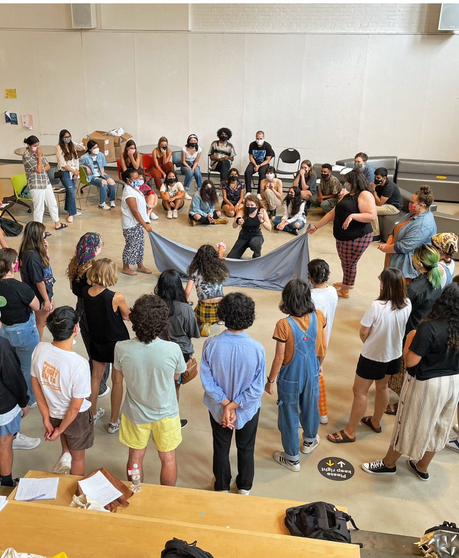 A group of students stands in a large circle indoors, participating in a group activity involving a large piece of fabric held in the center. The students are engaged and focused, with some sitting and others standing around the circle. Many of them are wearing masks, indicating a collective and inclusive environment.