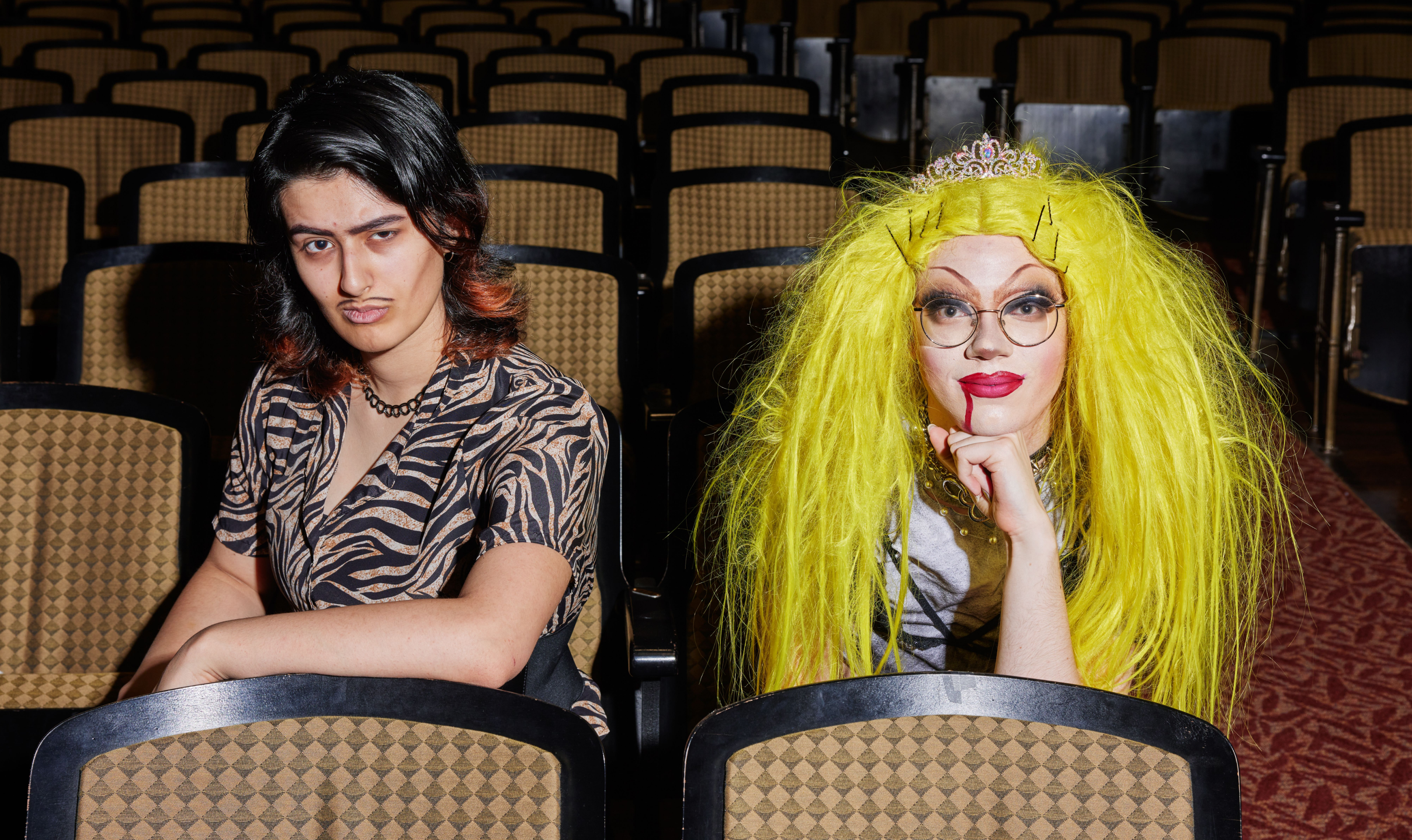 Two individuals are seated in a theater with rows of empty seats behind them. The person on the left has a neutral expression, dressed in a zebra-print shirt, while the person on the right, with a bright yellow wig and glasses, leans forward with a more playful demeanor. This image highlights diversity and self-expression.