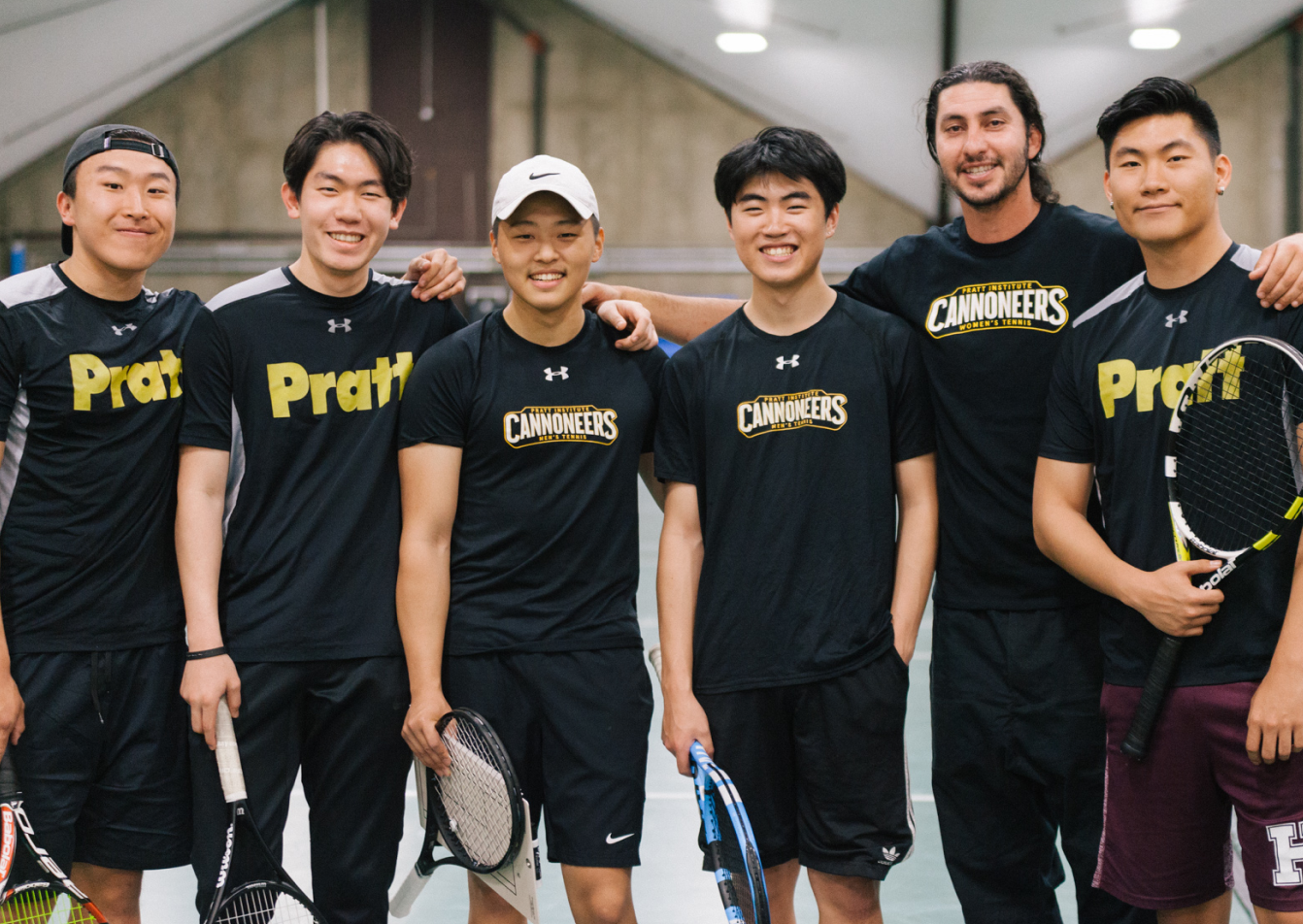 A group of six young men in athletic gear, likely members of a tennis team, stand together indoors, smiling for the camera. They are wearing matching black shirts with