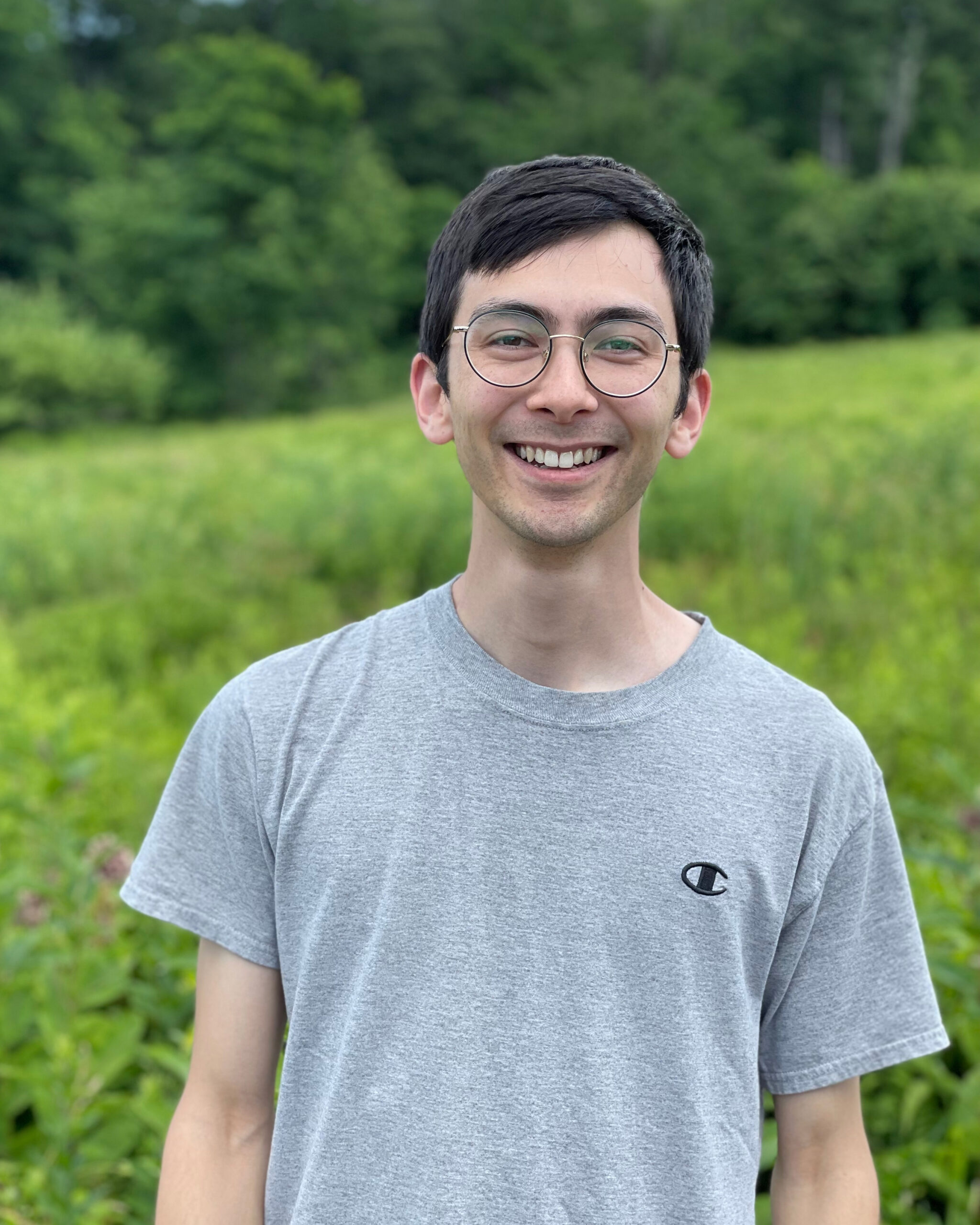 Headshot of Benjamin Goulet-Scott smiling, wearing grey Champion t-shirt, with a large open field and forest behind him, torso-length image