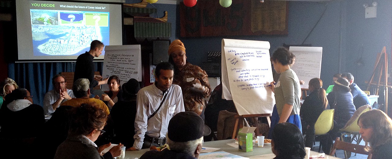 People sit at different roundtables in a room with slide show in the background. Each table has white board where they are writing for their respective groups.