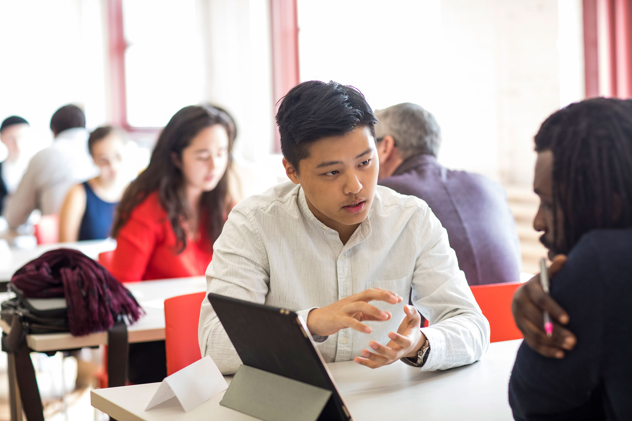 Two men are in a conversation together sitting at a table. An iPad is set between them. There are other colleagues at the table behind them.