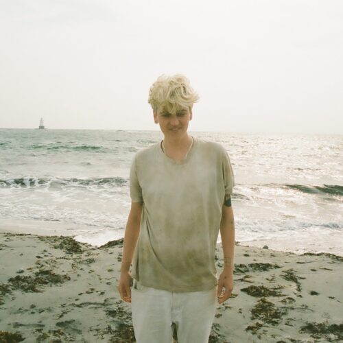 Tess Brown stands on a beach with her back facing towards the ocean. She is wearing white pants and a grey t-shirt