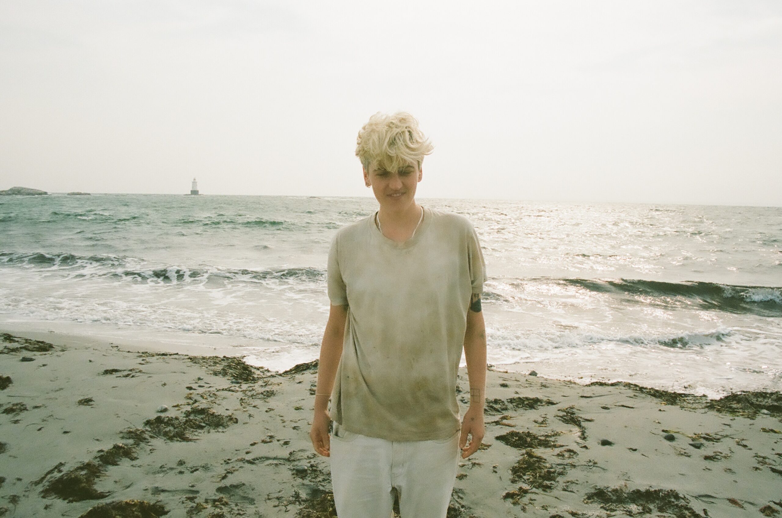 Tess Brown stands on a beach with her back facing towards the ocean. She is wearing white pants and a grey t-shirt