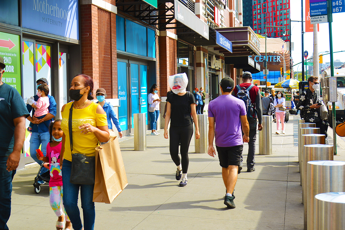 picture of urban sidewalk landscape, with groups of people walking up and down it, in brooklyn, barclays center behind them