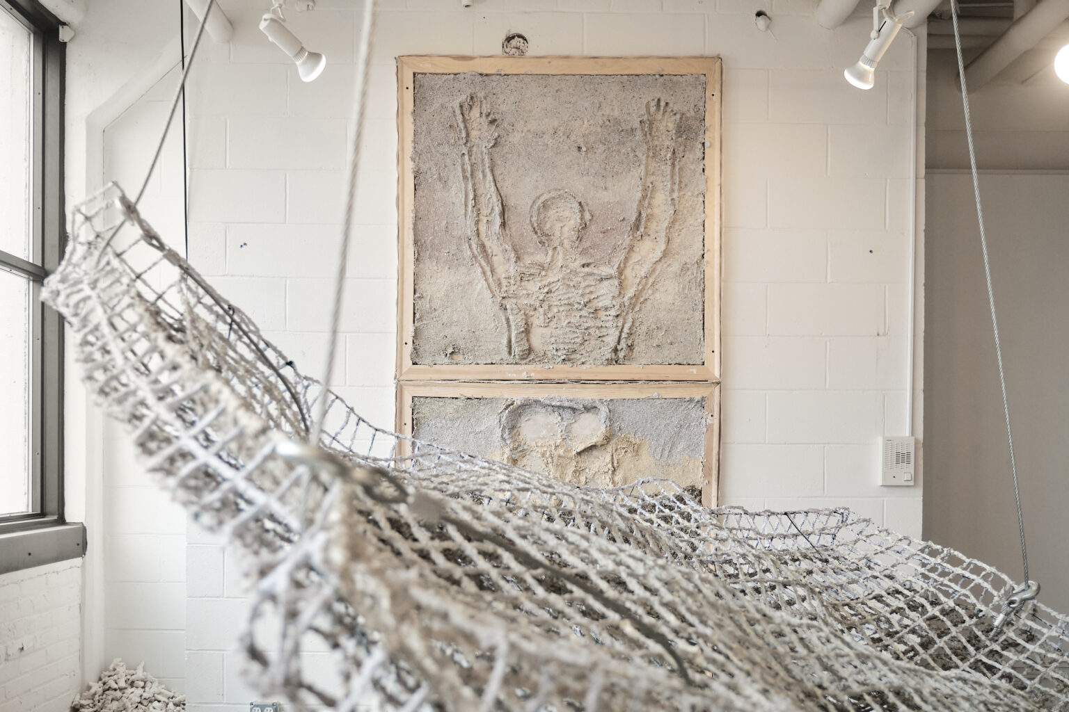 Sculpture consisting of gridded hammock like structure, with concrete panels on wall, with drawing of a person with their arms raised