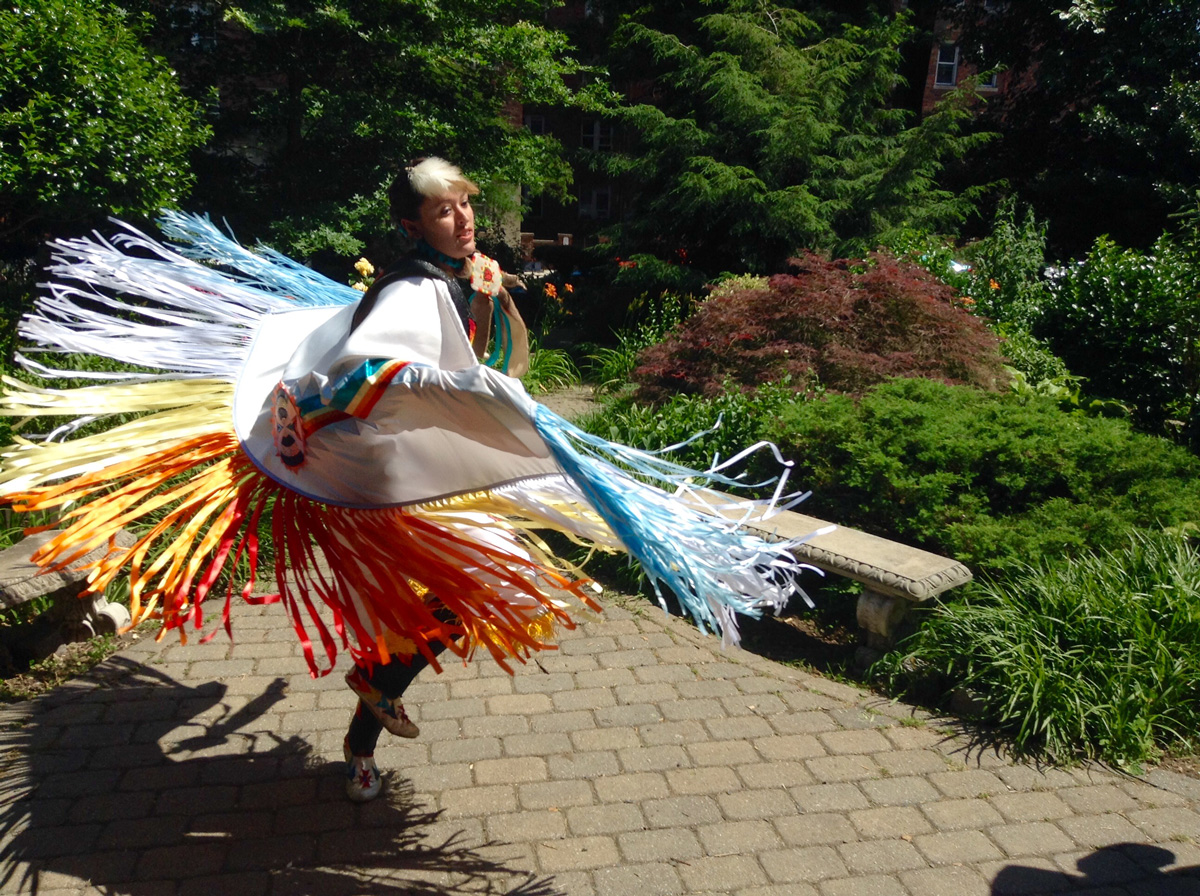 Dancer twirling in cape with long multicolored fringe, on sun-drenched brick path surrounded by lush greenery