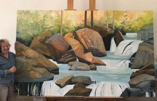 An artist stands next to a large painting of a gray-blue stream flowing through and over brown and dark gray boulders