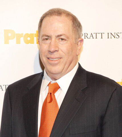 Headshot of a Bruce Gitlin. He is wearing a tuxedo with an orange tie. He is smiling.