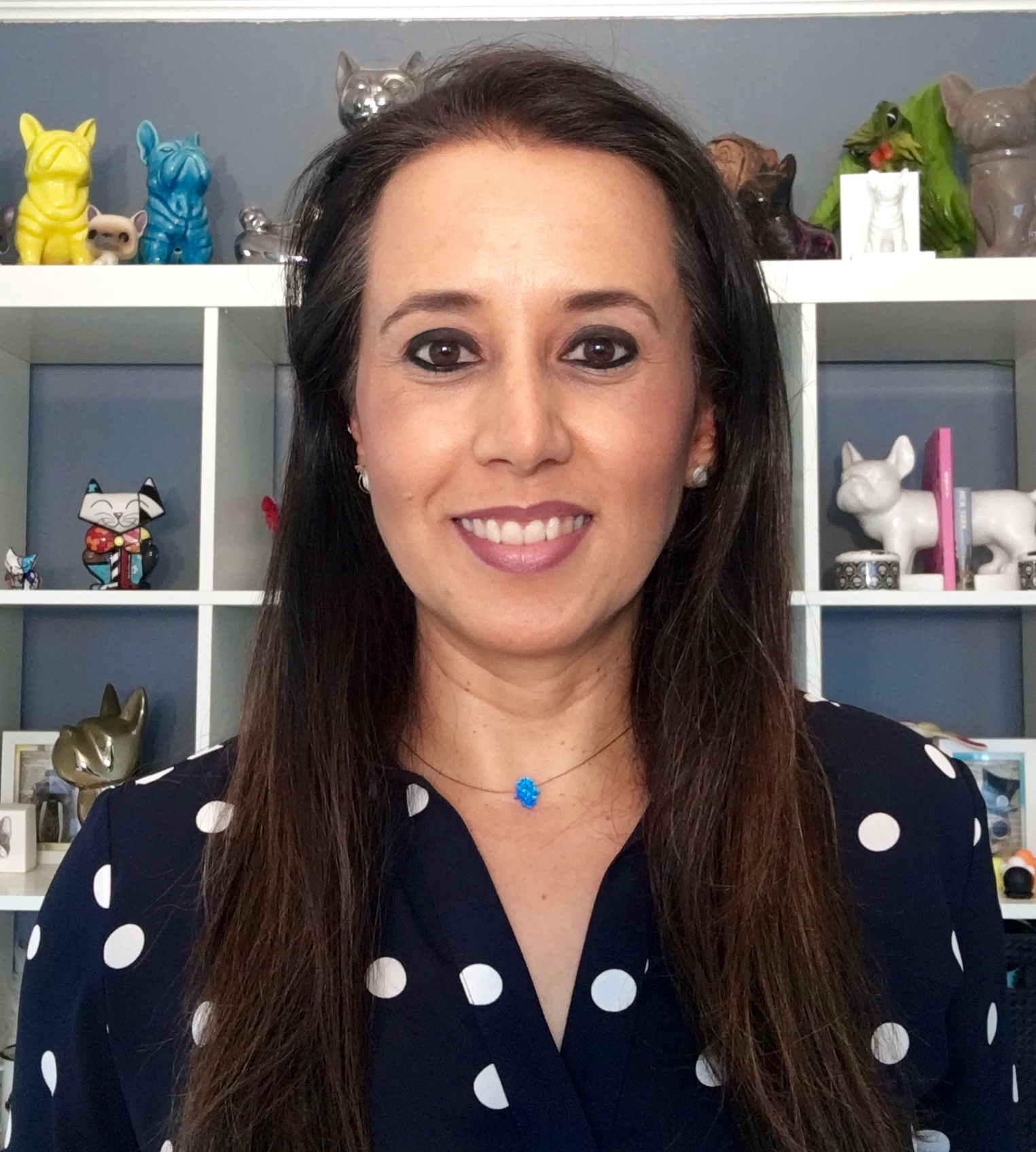 Headshot of Graciella Carillo, with long dark brown hair, brown eyes, and black-and-white polka-dot shirt against a bookcase full of whimsical sculptures of cats and dogs