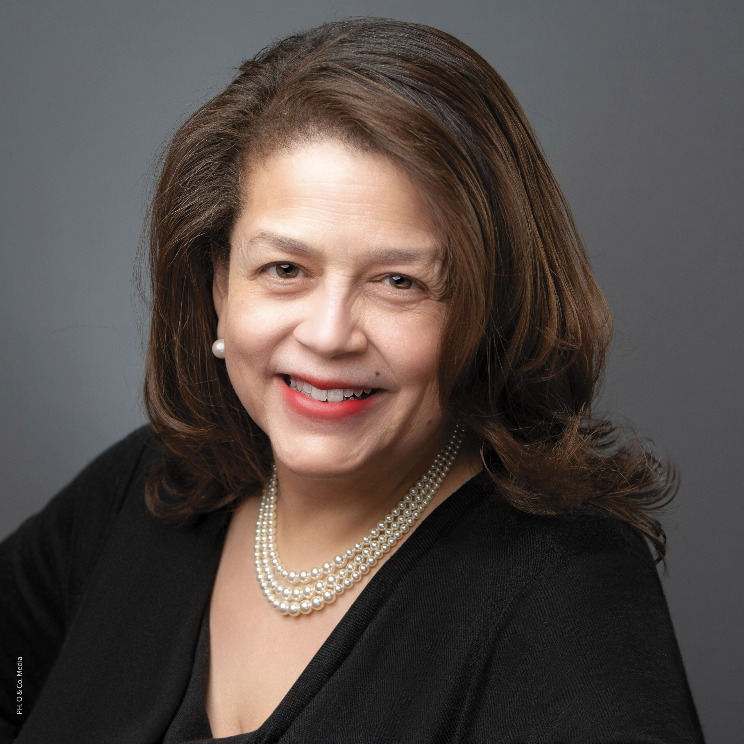 Cheryl D. Miller, smiling with shoulder length chestnut brown hair curled at the ends, brown eyes, and pearl necklaces and earrings