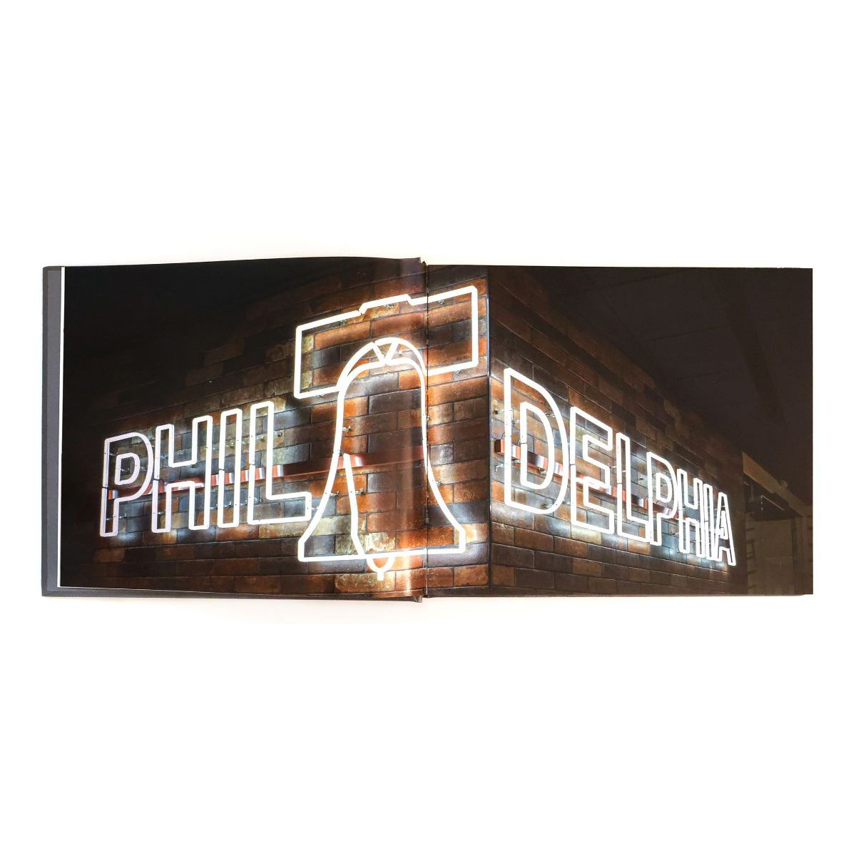 Book open to a spread showing the word Philadelphia shaped out of neon lights in block letters, with a silhouette of the Liberty Bell standing in for the first letter A, against a brick backdrop