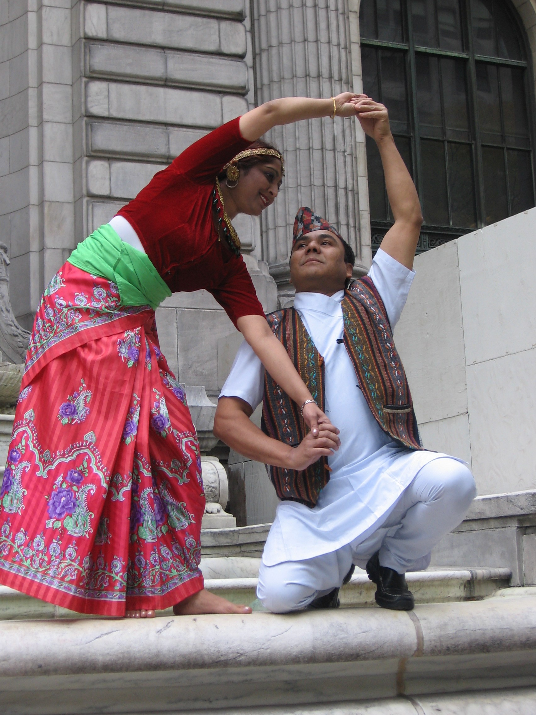 Dancer in a red skirt with purple and green flourishes, a deep red top, and golden headband arches over a second, kneeling dancer in white with a patterned vest perched on a marble step