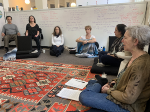 A group of people meditate in a room while sitting on the floor. There is a colorful rug in the center of the room